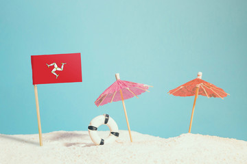 Miniature flag of Isle Of Man on beach with colorful umbrellas and life preserver. Travel concept, summer theme.
