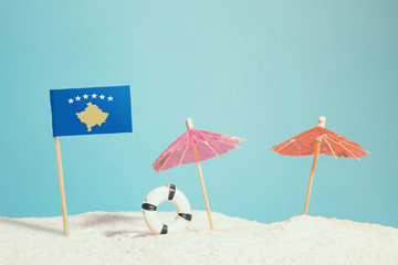 Miniature flag of Kosovo on beach with colorful umbrellas and life preserver. Travel concept, summer theme.