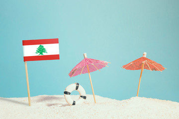 Miniature flag of Lebanon on beach with colorful umbrellas and life preserver. Travel concept, summer theme.