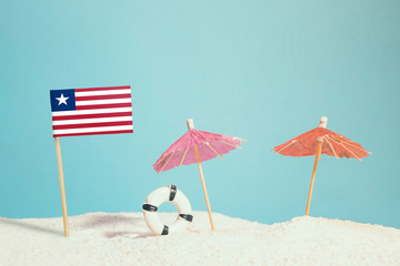 Miniature flag of Liberia on beach with colorful umbrellas and life preserver. Travel concept, summer theme.