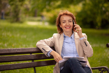 one smiling woman, looking at camera, while talking over phone. sitting in park on a bench.