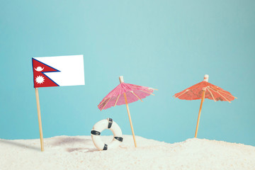 Miniature flag of Nepal on beach with colorful umbrellas and life preserver. Travel concept, summer theme.