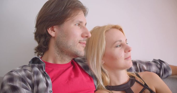 Closeup portrait of adult affectionate caucasian couple hugging tenderly smiling happily sitting on sofa indoors in cozy apartment