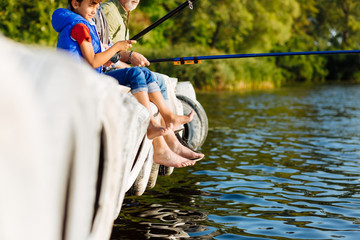 Boy sitting near father and grandfather having jeans pulled up while fishing