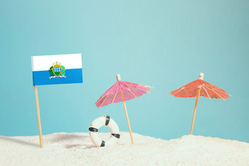 Miniature flag of San Marino on beach with colorful umbrellas and life preserver. Travel concept, summer theme.