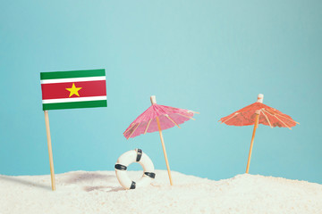 Miniature flag of Suriname on beach with colorful umbrellas and life preserver. Travel concept, summer theme.
