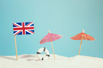Miniature flag of United Kingdom on beach with colorful umbrellas and life preserver. Travel concept, summer theme.