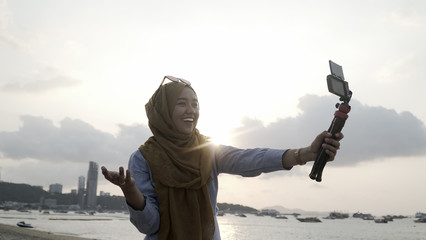 Blogger Asian Muslim woman record vlog video on beach, young beautiful female happy to using phone making video vlog