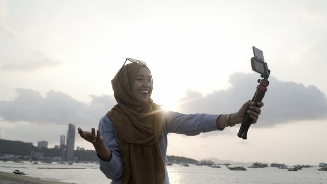 Blogger Asian Muslim woman record vlog video on beach, young beautiful female happy to using phone making video vlog