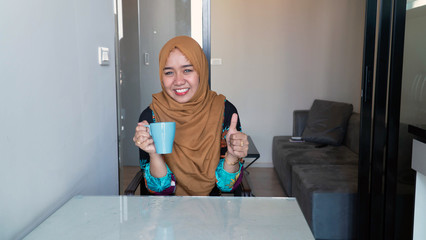 Fototapeta na wymiar portrait of Happy muslim woman with hijab sitting in kitchen holding cup of tea of coffee enjoying her relaxing free time