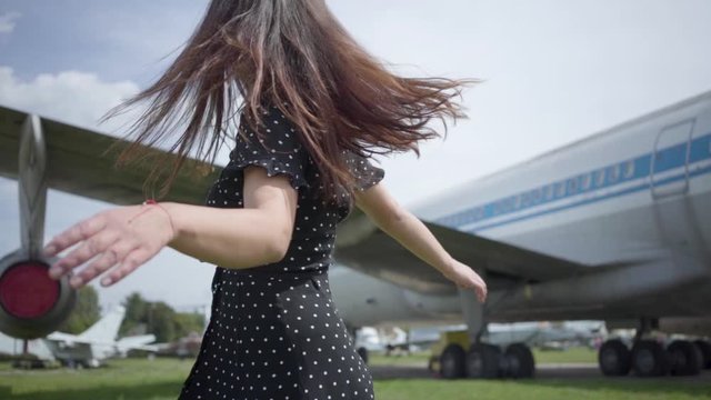 Beautiful girl walks next to the plane, turning and spinning, looking at the camera. Summertime. Joy of travel. Concept of traveling, aircraft, weekend. Slow motion.