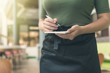 Cropped image of woman waitress in apron ready to take order using notepad and pen in cafe or...