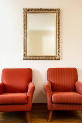 Two chairs and golden framed mirror on a white wall with soft natural light coming in from outside. beauty