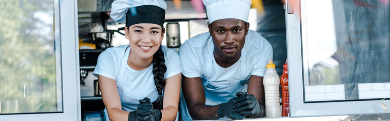 panoramic shot of asian girl and african american man in hats smiling from food truck