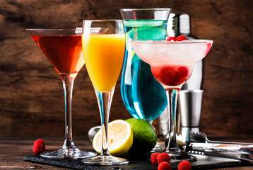 Colorful summer cocktails. Cold alcoholic beverages and drinks: mimosa, cosmopolitan, raspberry margarita and blue hawaii on wooden table background with bar tools