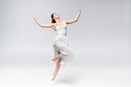 young elegant ballerina in white dress dancing on grey background