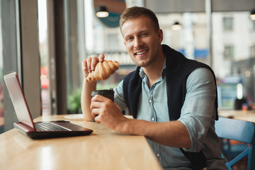 young handsome smiling man eating his croissant and drinking hot coffee for lunch in the cafe while working remotely in his laptop