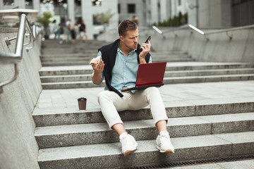 young handsome office worker having unpleasant business conversation while eating his sandwich and...