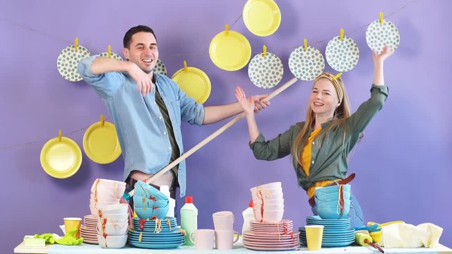 Young funny man and woman singing songs and dancing with cleaning equipment. Hanging clean plates in background of photo.