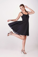 Naklejka premium Young attractive sexy blond woman model with light hair, wearing short black dress and sandals on heels, posing and dancing in white studio