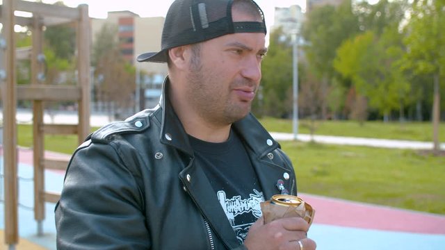 Portrait of a hooligan in the biker jacket telling something and illegally drinking beer on the kid's playground