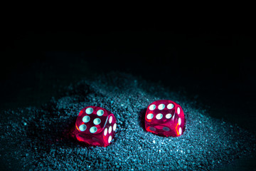 Bright red dice in the black sand. Evening time. Black background of a wooden Board.
