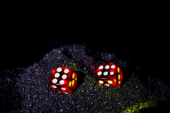 Bright red dice in the black sand. Evening time. Black background of a wooden Board.