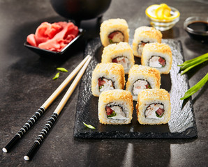 Macro shot of tempura uramaki sushi rolls with smoked chicken, cream cheese, green onion, tomato, rice and nori. Chopsticks holding hot crispy maki roll on black stone slate background closeup