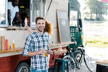 selective focus of happy man holding bottle with beer and carton plate with street food near woman and african american man in food truck