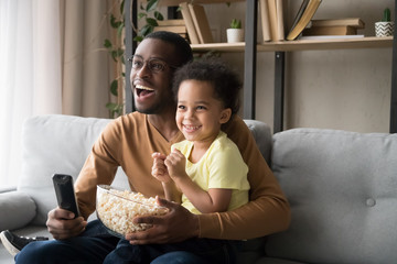 Happy African American father with son watching tv, eating popcorn