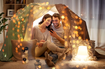 leisure, hygge, technology and people concept - happy couple with smartphone in kids tent at home