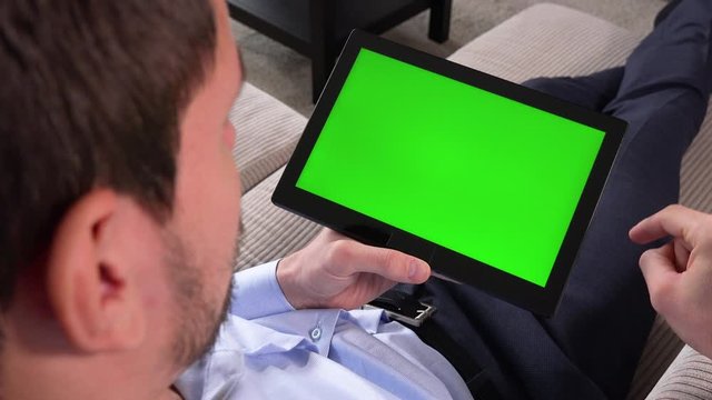 A man sits on a couch in an apartment and works on a tablet with a green screen in horizontal position - closeup - scrolls down and taps at something