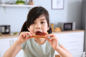 Funny little girl eating tasty toast with jam in kitchen