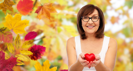 Obraz na płótnie Canvas health, charity and valentine's day concept - portrait of smiling senior woman holding red heart over autumn leaves and nature background