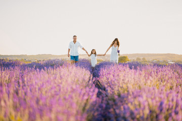 Happy Caucasian Family in Purple Lavender Field. Parent Couple and Little Girl Enjoy Walking Blooming Meadow on Summer Day. Beautiful Mom and Handsome Dad Holding Pretty Daughter Hand Together