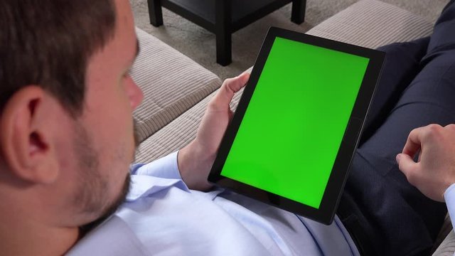 A man sits on a couch in an apartment and works on a tablet with a green screen in vertical position - closeup - he zooms in and out
