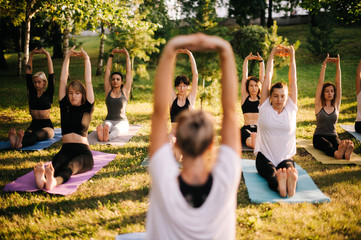 Group of young women are practicing yoga and sits with hands up morning in park while sunrise....