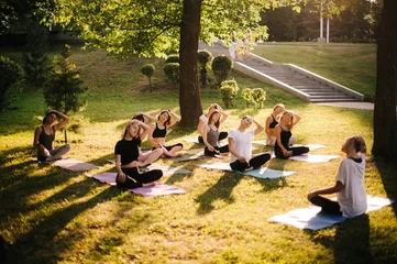 Fotobehang Ontspanning Group of women are stretching neck in city park on summer sunny morning under guidance of instructor. Group of people outdoors is sitting on yoga mat in lotus pose on grass with eyes closed.  © dikushin