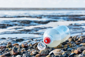 Plastic bottle with red bottle cap on a stony beach with shells at low tide. Concept of plastic pollution of the sea and marine life. Beat Plastic Pollution. Blue sky with clouds and copy space.