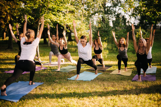 Group of young sporty handsome women practicing yoga lesson with instructor in city park on summer sunny morning. Group of people are standing together in Virabhadrasana 1 exercise, Warrior one pose