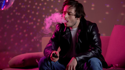 Idle young man vaping e-cigarette to relieve stress, relaxing at nightclub party