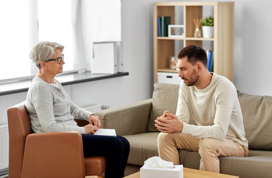 psychology, mental therapy and people concept - senior woman psychologist talking to sad young man patient at psychotherapy session