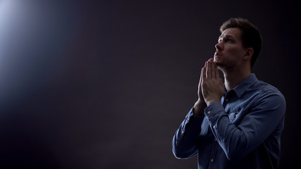 Young man praying in dark room under blessed light from heaven, Christianity