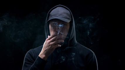 Male bully in hoodie smoking cigarette against dark background, night criminal
