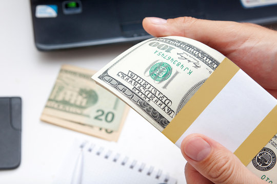 hundreds of US dollars in a stack holds a male hand on the background of a desktop with a laptop - concept photo of business and ideas for making money