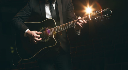 Fototapeta na wymiar Man in jacket plays and sings while standing on stage on a 12-string acoustic guitar. Dark background and blurred back light