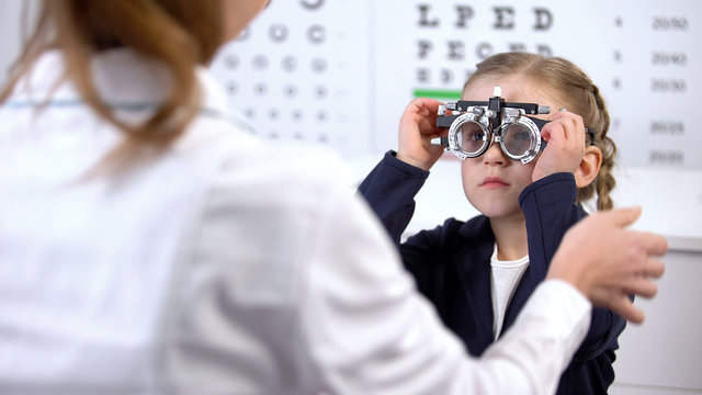 Pretty little schoolgirl visiting oculist for measuring eyesight with phoropter