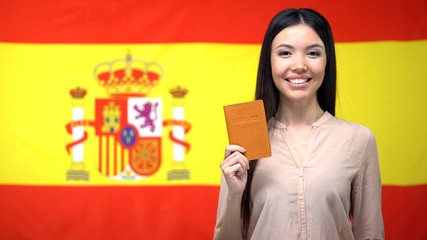 Smiling Asian girl holding passport against Spanish flag background, citizenship
