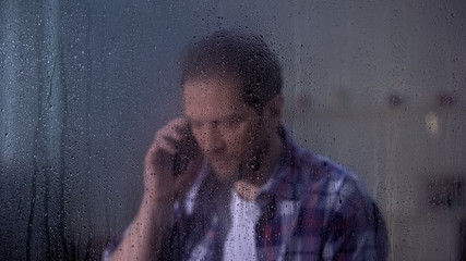 Middle-aged male talking phone with family behind rainy window missing relatives