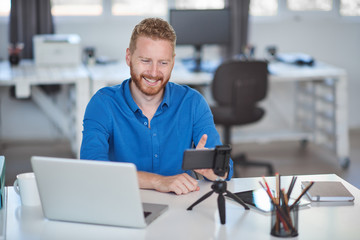 Young Caucasian employee having a video call over smart phone while sitting in office. On desk is laptop. Start up business concept.